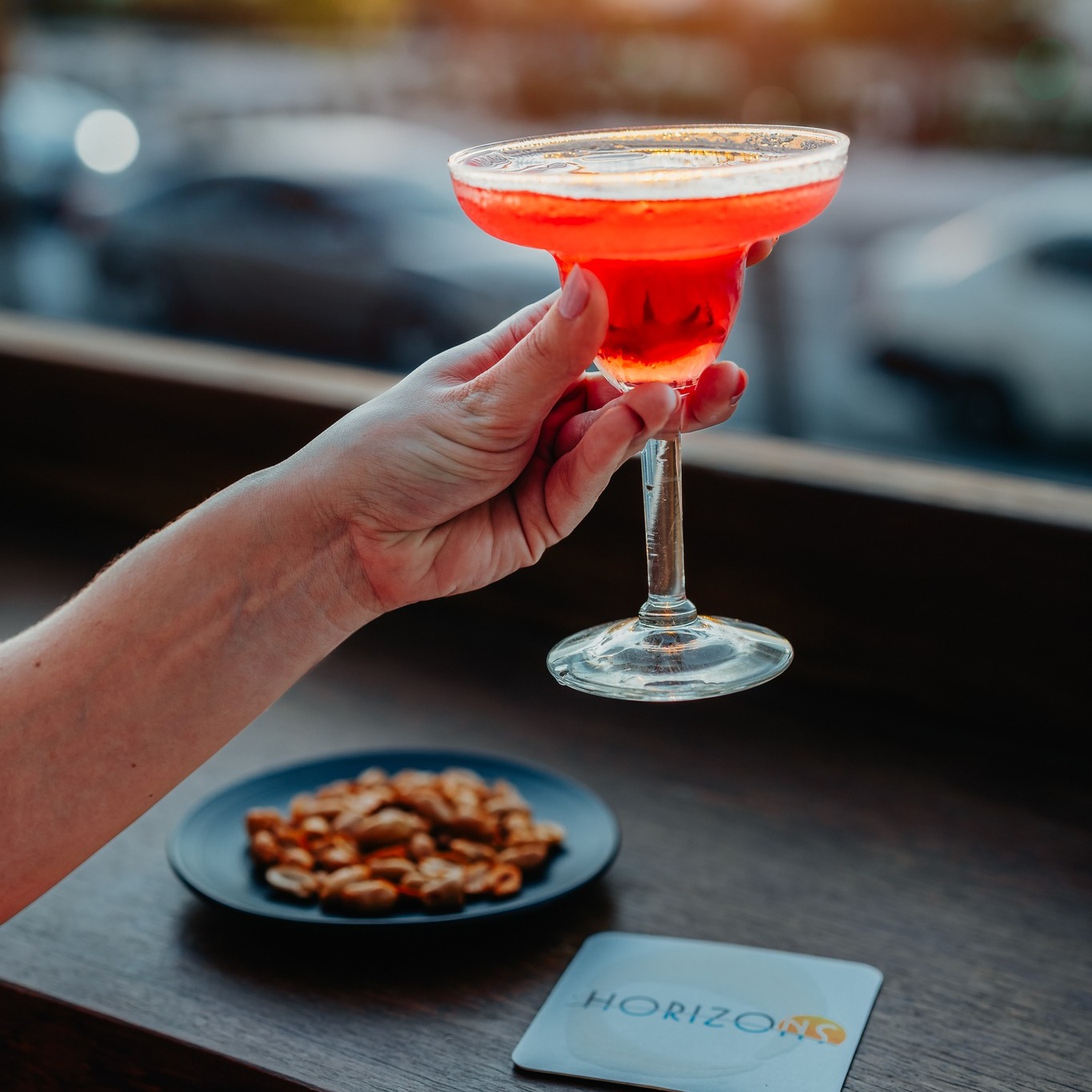 Hand raising a red signature cocktail over a plate of appetizers and Horizons branding
